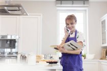 Female caterer with cookbook baking, talking on cell phone in kitchen — Stock Photo