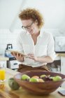 Портрет женщины, сидящей и смотрящей на цифровой планшет на кухне, улыбающейся — стоковое фото