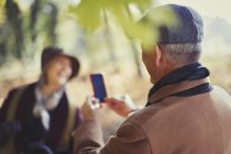 Uomo anziano con fotocamera telefono fotografare moglie nel parco — Foto stock