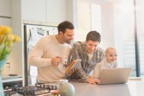 Male gay parents with baby son using digital tablet and laptop in kitchen — Stock Photo