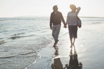 Affectueux couple d'âge mûr tenant la main et marchant sur le coucher du soleil océan plage surf — Photo de stock