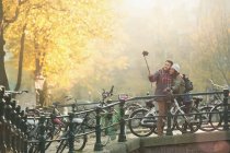 Pareja joven con bicicletas tomando selfie con palo de selfie en el puente de otoño, Amsterdam - foto de stock
