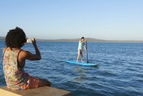 Joven fotografiando amigo paddleboarding en el soleado océano de verano - foto de stock