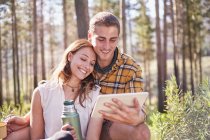 Young couple camping, drinking coffee and using digital tablet in sunny woods — Stock Photo