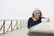 Усміхнена старша жінка бере селфі з телефоном на пляжній дошці — стокове фото