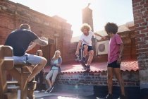 Homem com câmera telefone fotografar amigo fazendo acrobacia skate em ensolarado telhado urbano — Fotografia de Stock