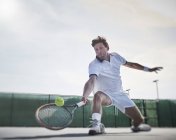 Determinado jovem jogador de tênis masculino jogando tênis, alcançando a bola na quadra de tênis ensolarada — Fotografia de Stock