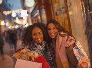 Retrato sonriente jóvenes amigas con bolsas de compras en la calle nocturna urbana - foto de stock