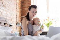 Mother holding daughter and working at laptop on bed — Stock Photo