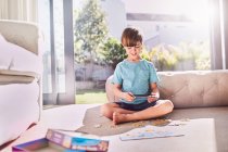 Junge mit digitalem Tablet setzt Puzzle auf sonnigem Wohnzimmerboden zusammen — Stockfoto