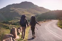 Young couple with backpacks hiking along sunny, remote roadside — Stock Photo