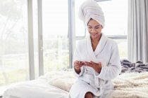Улыбающаяся женщина в халате и завернутые в полотенце волосы с помощью цифрового планшета на кровати — стоковое фото