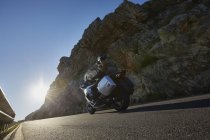 Couple riding motorcycle on sunny road along craggy cliff — Stock Photo