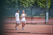 Jeune couple de joueurs de tennis marchant avec des raquettes de tennis sur un court de tennis en terre battue ensoleillé — Photo de stock