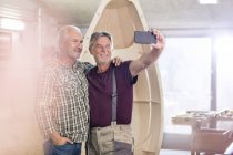 Гордый, улыбающийся мужчина-плотник с камерой телефона делает селфи рядом с деревянной лодкой в мастерской — стоковое фото