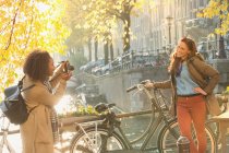 Young woman photographing friend with bicycle along autumn canal, Amsterdam — Stock Photo