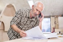 Senior carpenter talking on cell phone and reviewing paperwork in workshop — Stock Photo