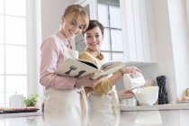 Female caterers with cookbook baking in kitchen, using electric hand mixer — Stock Photo