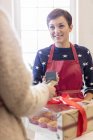 Female caterer selling box of baked pastries to woman using smart phone credit card reader — Stock Photo