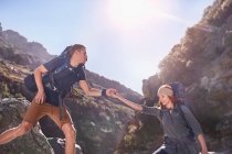 Young man helping girlfriend hiking below sunny, craggy cliffs — Stock Photo