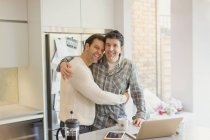 Portrait affectionate male gay couple hugging at laptop in kitchen — Stock Photo