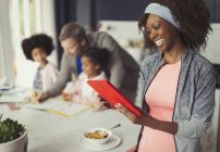 Smiling mother using digital tablet and eating breakfast in kitchen with young family — Stock Photo