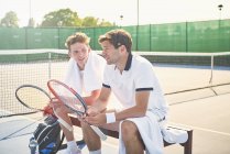 Junge männliche Tennisspieler ruhen sich mit Tennisschlägern auf sonnigem Tennisplatz aus — Stockfoto