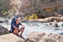 Young man with backpack hiking, resting at sunny, craggy stream — Stock Photo