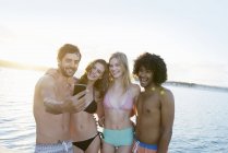 Young adult friends in bikinis and swim trunks taking selfie at summer sunset ocean — Stock Photo
