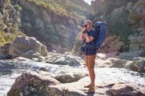Young man with backpack hiking, photographing with camera on sunny rocks — Stock Photo