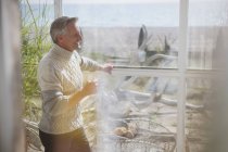 Mature man in turtleneck sweater drinking coffee in sunny beach house sun porch — Stock Photo