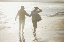 Mature couple holding hands and walking on sunny beach — Stock Photo