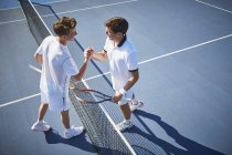 Молодые теннисисты пожимают руку в теннисной сетке на солнечном синем теннисном корте — стоковое фото