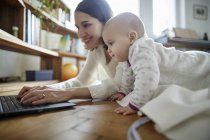 Baby daughter watching mother typing on laptop on floor — Stock Photo
