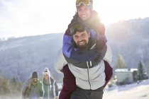 Retrato brincalhão casal piggybacking em ensolarado, campo nevado — Fotografia de Stock
