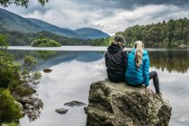 Pareja sentada en una roca mirando un lago tranquilo, Loch an Eilein, Escocia - foto de stock