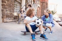 Couple souriant avec ballon de football et planche à roulettes parlant sur le trottoir urbain — Photo de stock