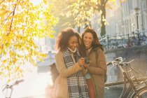 Smiling young women friends with digital camera along sunny urban autumn canal, Amsterdam — Stock Photo