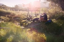 Casal jovem com mochilas caminhadas, descansando em campo remoto ensolarado — Fotografia de Stock