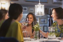 Mujeres sonrientes amigas brindando por copas de vino blanco cenando en la mesa del restaurante - foto de stock