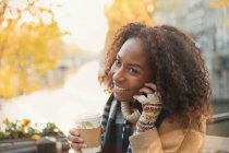 Smiling young woman drinking coffee and talking on cell phone at autumn sidewalk cafe — Stock Photo