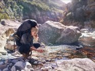Jeune femme avec sac à dos randonnée, se laver les mains au ruisseau ensoleillé — Photo de stock