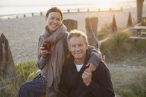 Retrato sonriente pareja madura cogida de la mano y beber vino en la playa puesta del sol - foto de stock