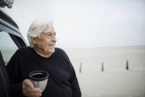 Happy senior man drinking coffee at car on winter beach — Stock Photo