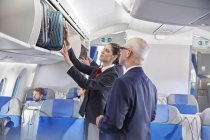 Asistente de vuelo ayudando a los hombres de negocios a colocar el equipaje en el compartimento superior en el avión - foto de stock