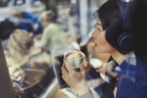Mujer joven pensativa escuchando música con auriculares y tomando café en la ventana de la cafetería - foto de stock