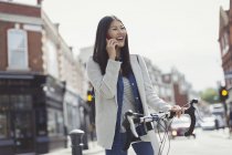 Улыбающаяся молодая женщина ездит на велосипеде, разговаривает по мобильному телефону на солнечной городской улице — стоковое фото