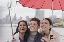 Smiling friend tourists with umbrella taking selfie with selfie stick, London, UK — Stock Photo