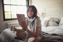 Усміхнена молода жінка з навушниками п'є каву і використовує цифровий планшет на ліжку — стокове фото