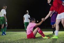 Young female soccer player helping fallen teammate get up on field at night — Stock Photo